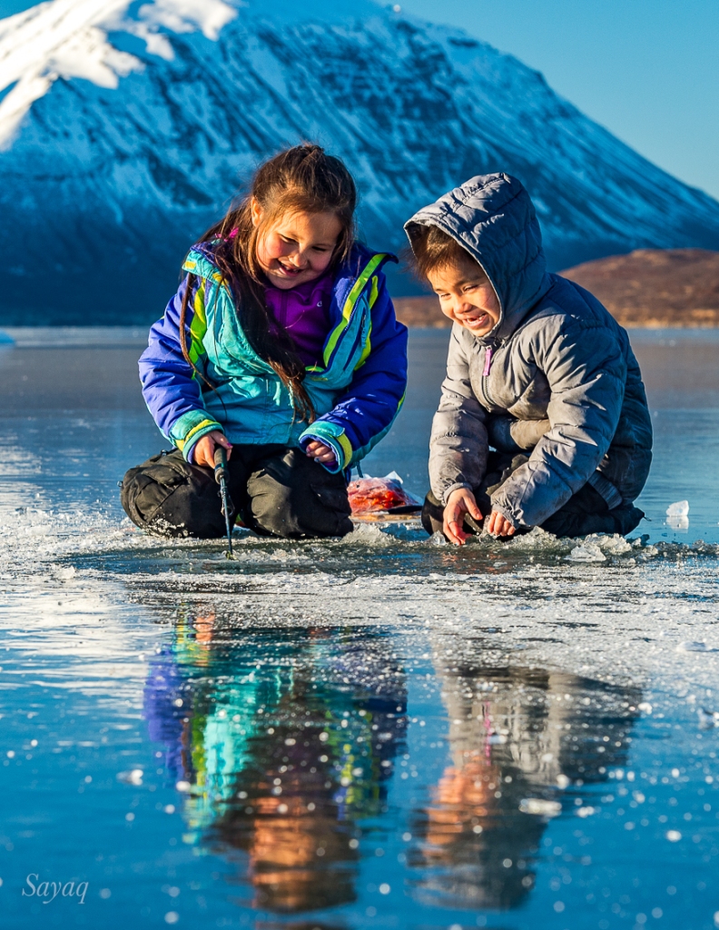 Photograph of Native Alutiiq girls colorfully reflected on lake ice as they jig for fish on frozen Chignik Lake, Alaska. 